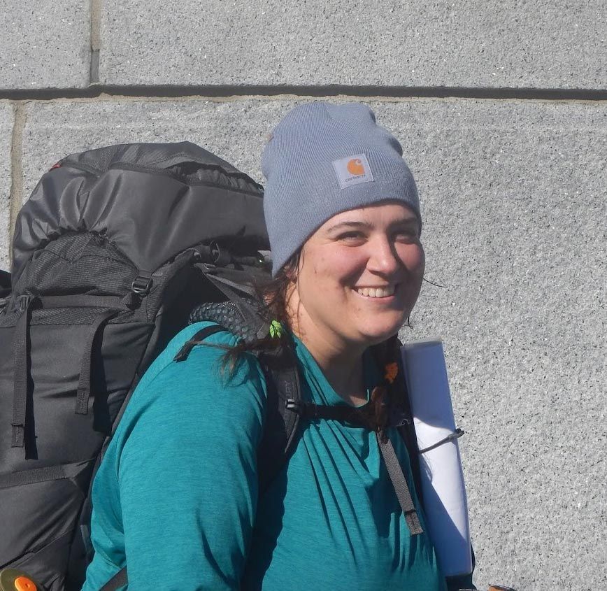 Eva Moynihan wearing a beanie and a large backpack, smiles at the camera.