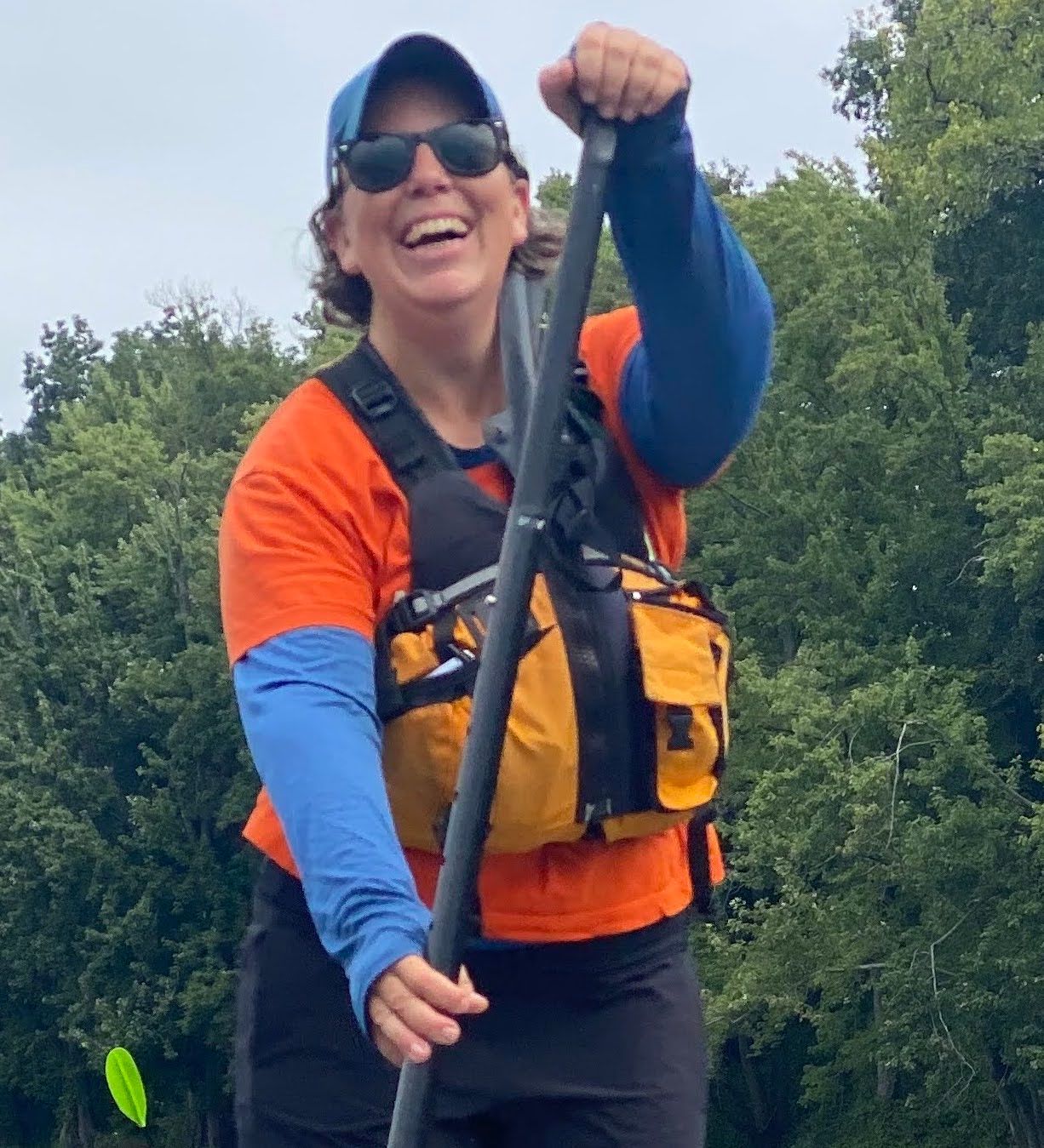 Karen Foster smiles while paddling a stand-up paddleboard in a hat, sunglasses, and other gear.