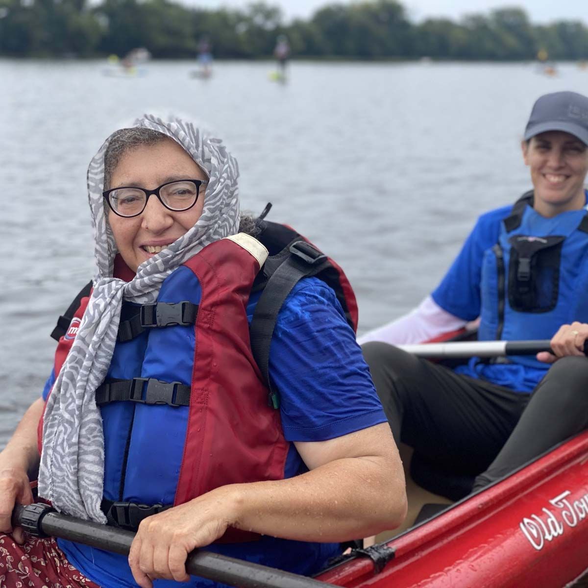 A woman with gray hair and glasses smiles at the camera from her seat in a kayak, on a river.