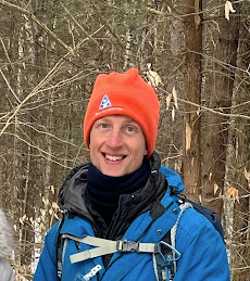 Kurt Kauppinen, in an All Out Adventures winter hat, smiles while hiking in the woods in winter.