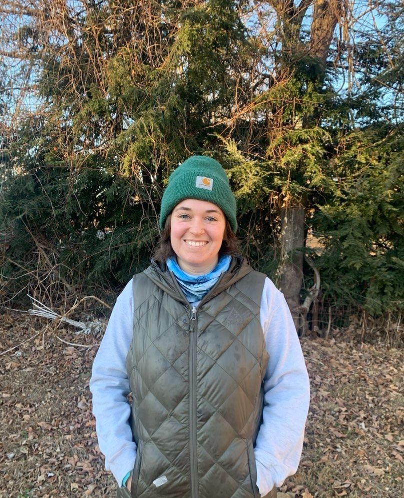 Molly Aronson, wearing a green beanie and a green vest, smiles at the camera in an outdoor setting, in front of trees.