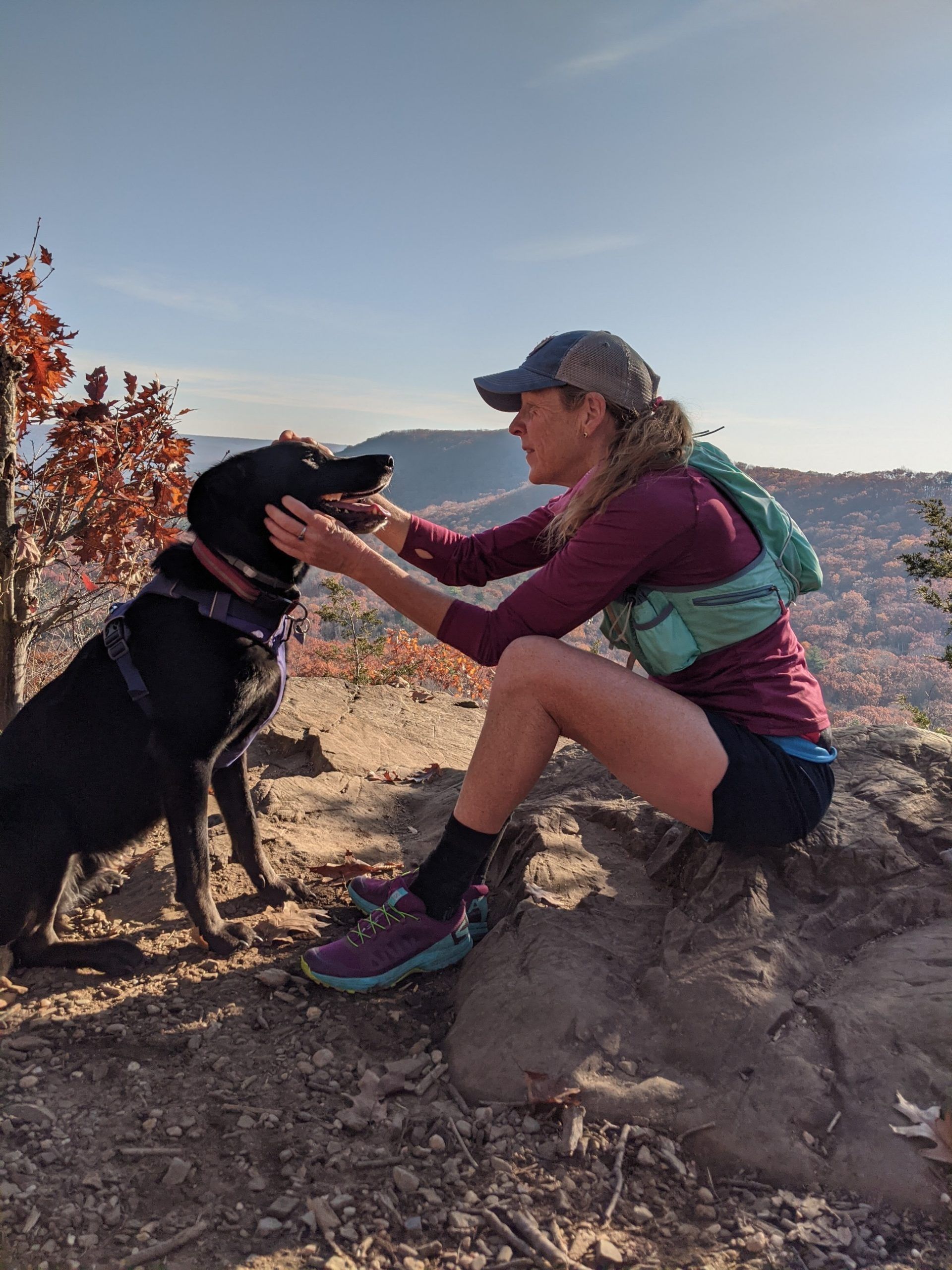 Patti Dougherty pets her dog while sitting on a rock, out hiking in a hilly area