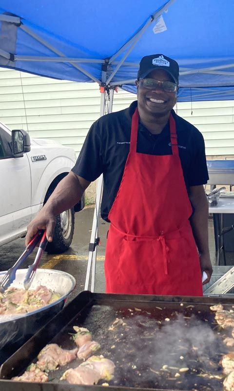 A man in a red apron is smiling and standing in front of a grill, cooking food.