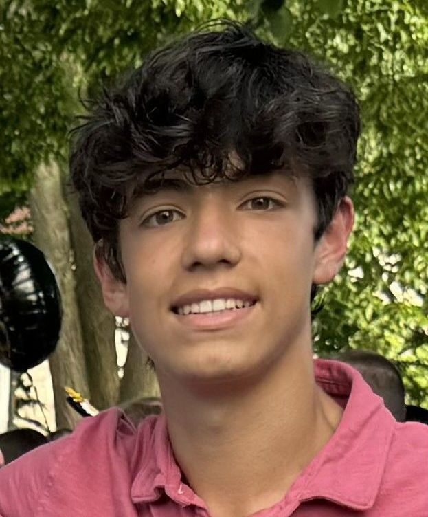 Image shows a young man in a pink collared shirt smiling