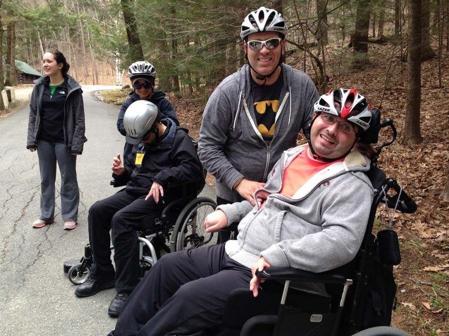 Four hikers, two in wheelchairs, are paused along a paved accessible trail through the woods. Two smile at the camera.