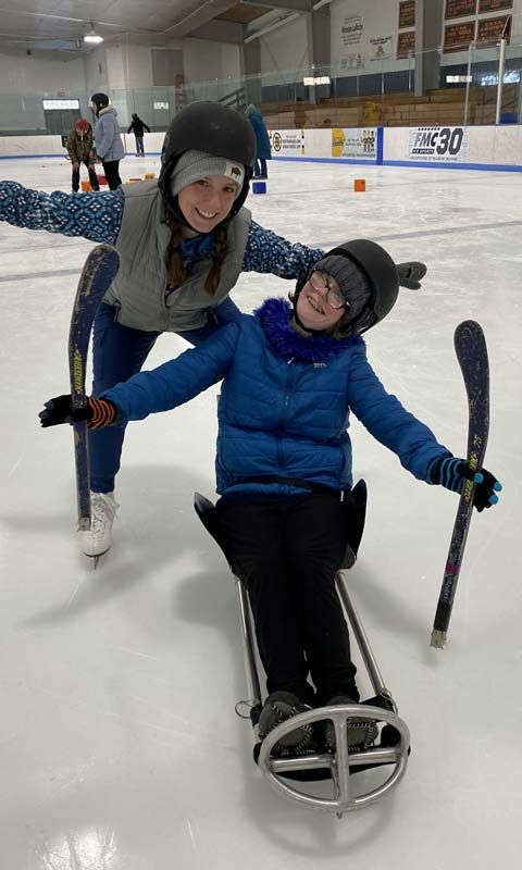 Two young people smile at the camera at an ice rink. One is on figure skates and the other is on a sit skating rig.