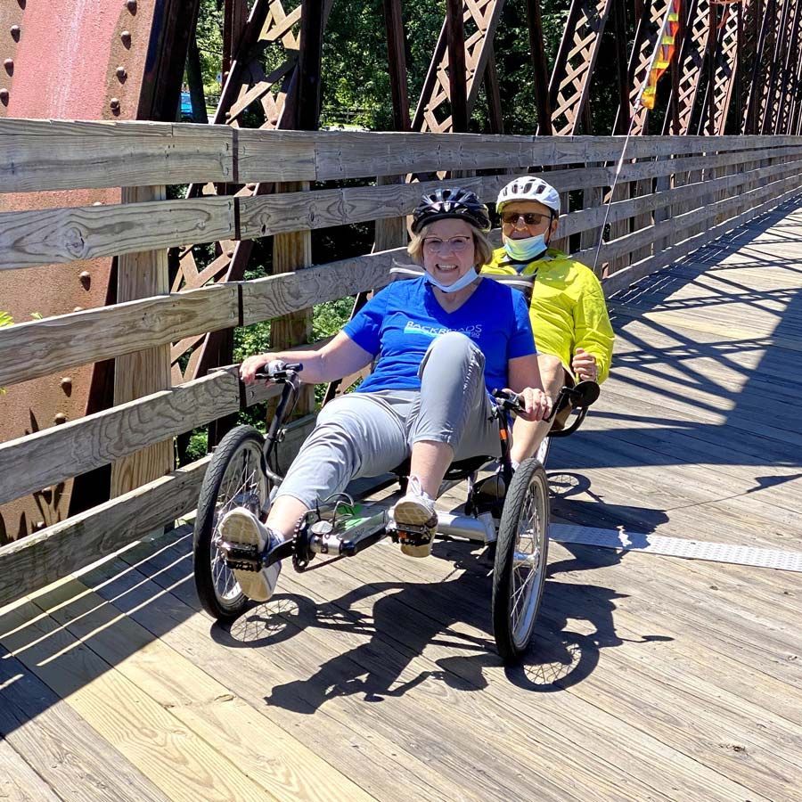 Two people enjoy riding a tandem recumbent bike on the Northampton rail trail bridge.