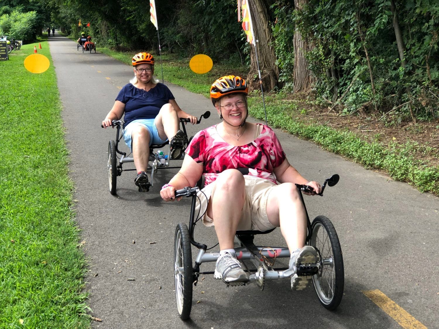 Two women in helmets smile as they ride recumbent tricycles on a bike path.