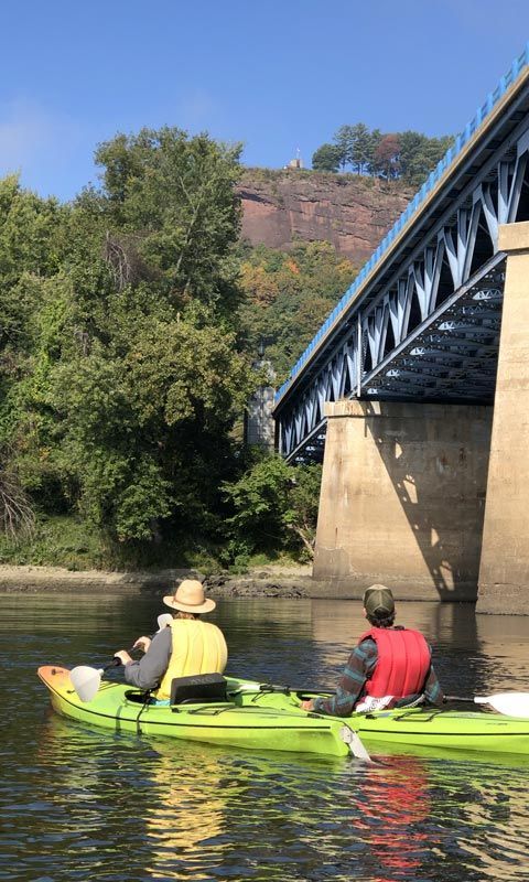 Two kayakers in lime green kayaks gaze up at a bridge and red rock cliff above a river.