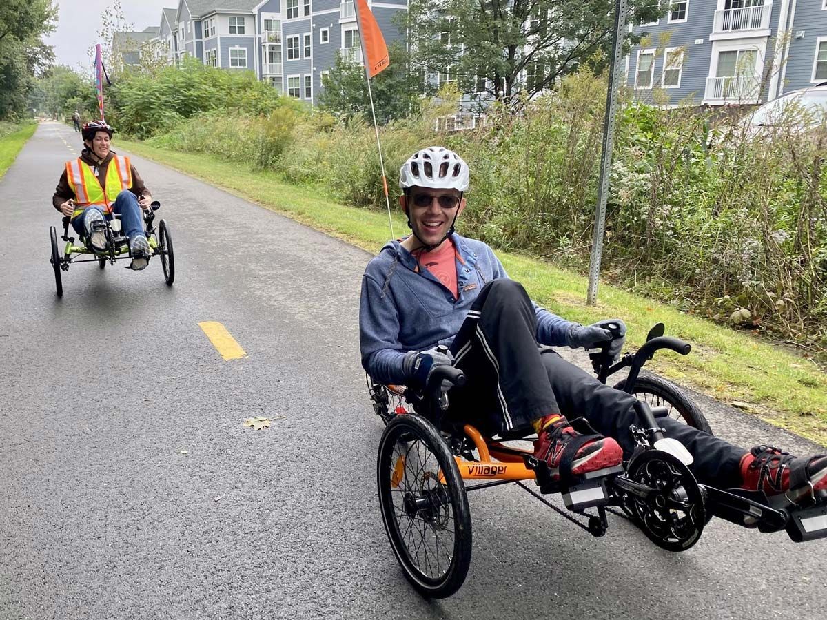 Two people are riding recumbent bicycles down a bike trail, smiling at the camera.