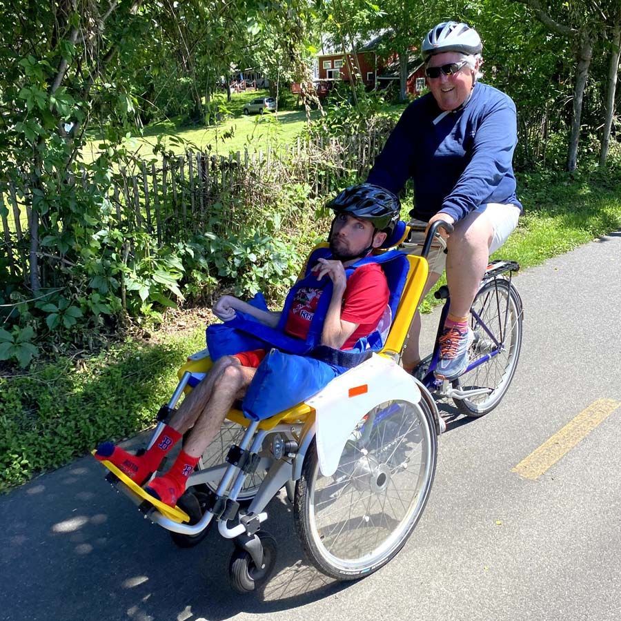 An able-bodied person and a young disabled man use a specialized wheelchair tandem cycle to enjoy a bike path.