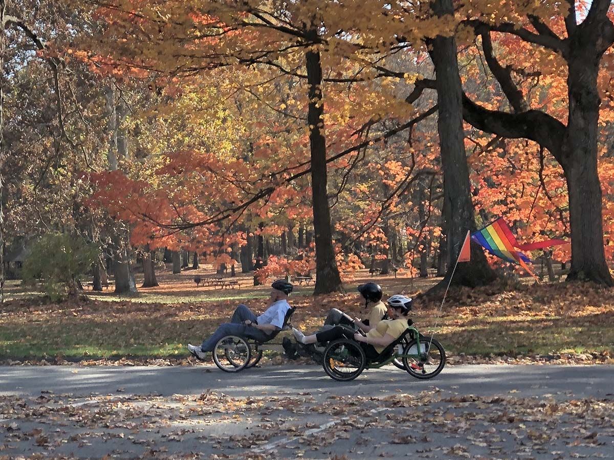 Three people traveling by recumbent tricycles on a paved path through colorful fall foliage.