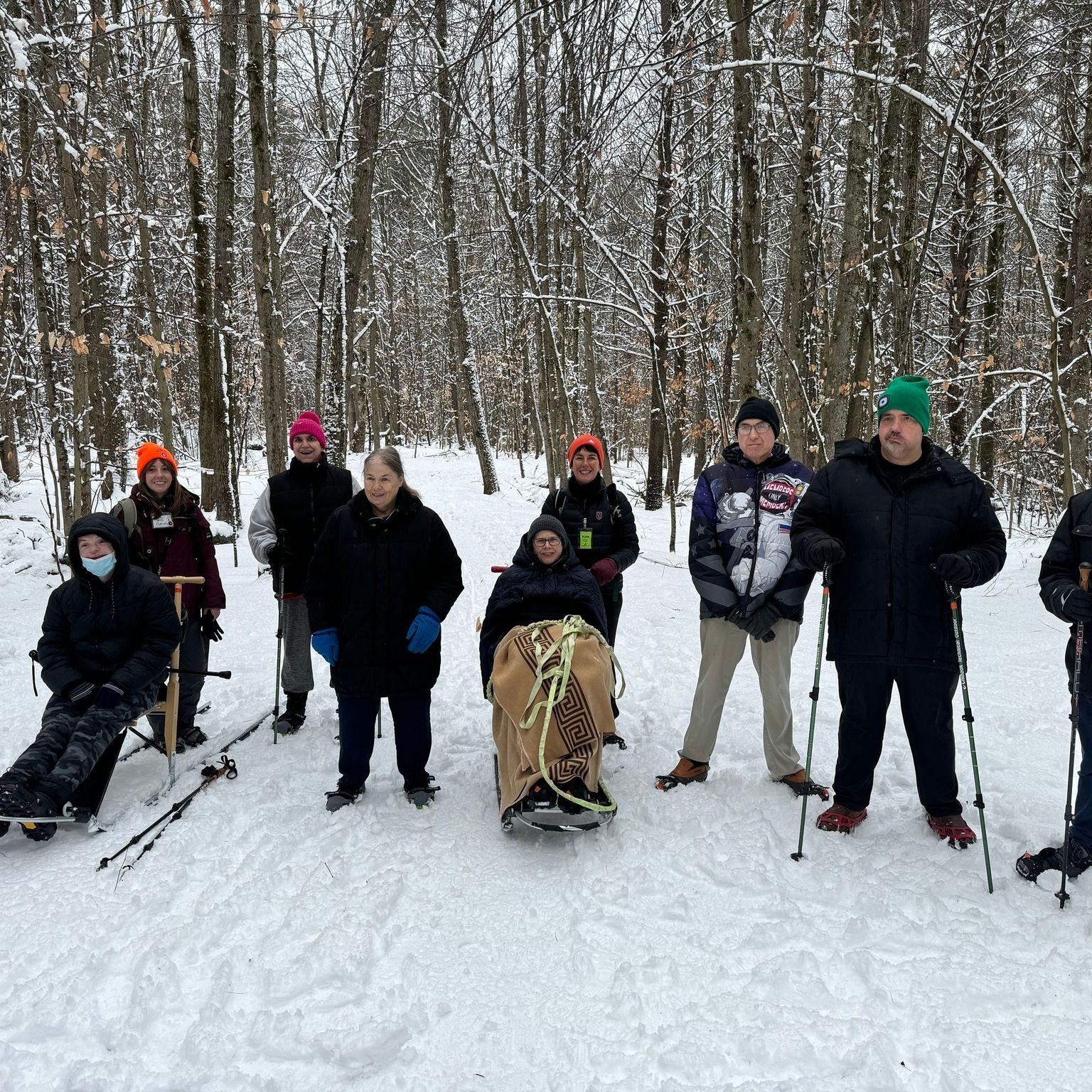 Image shows a group of people on a snowy hiking trail.  Several are standing with hiking poles, 2 are seated on kicksleds.