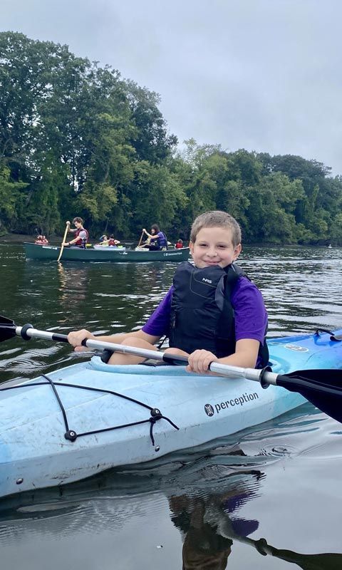 A young boy with a paddle in hand smiles at the camera from his kayak on a river.