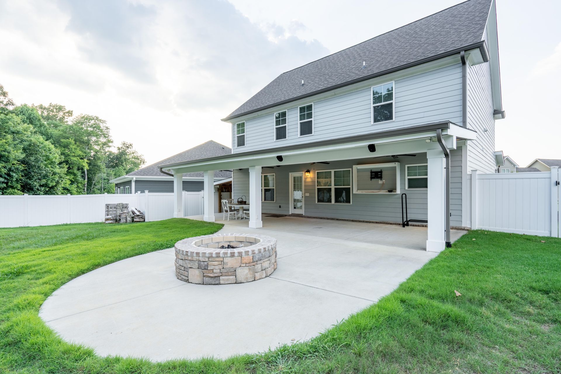 A backyard patio with a stone fire pit, covered porch, and white vinyl fence against a two-story gray house.