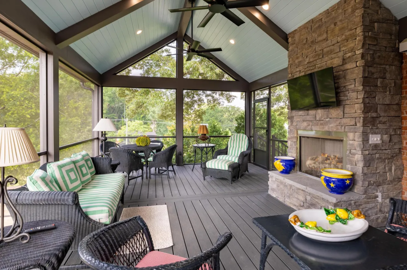 A screened-in porch with dark wicker furniture, a stone fireplace, and a light blue vaulted ceiling overlooking trees.