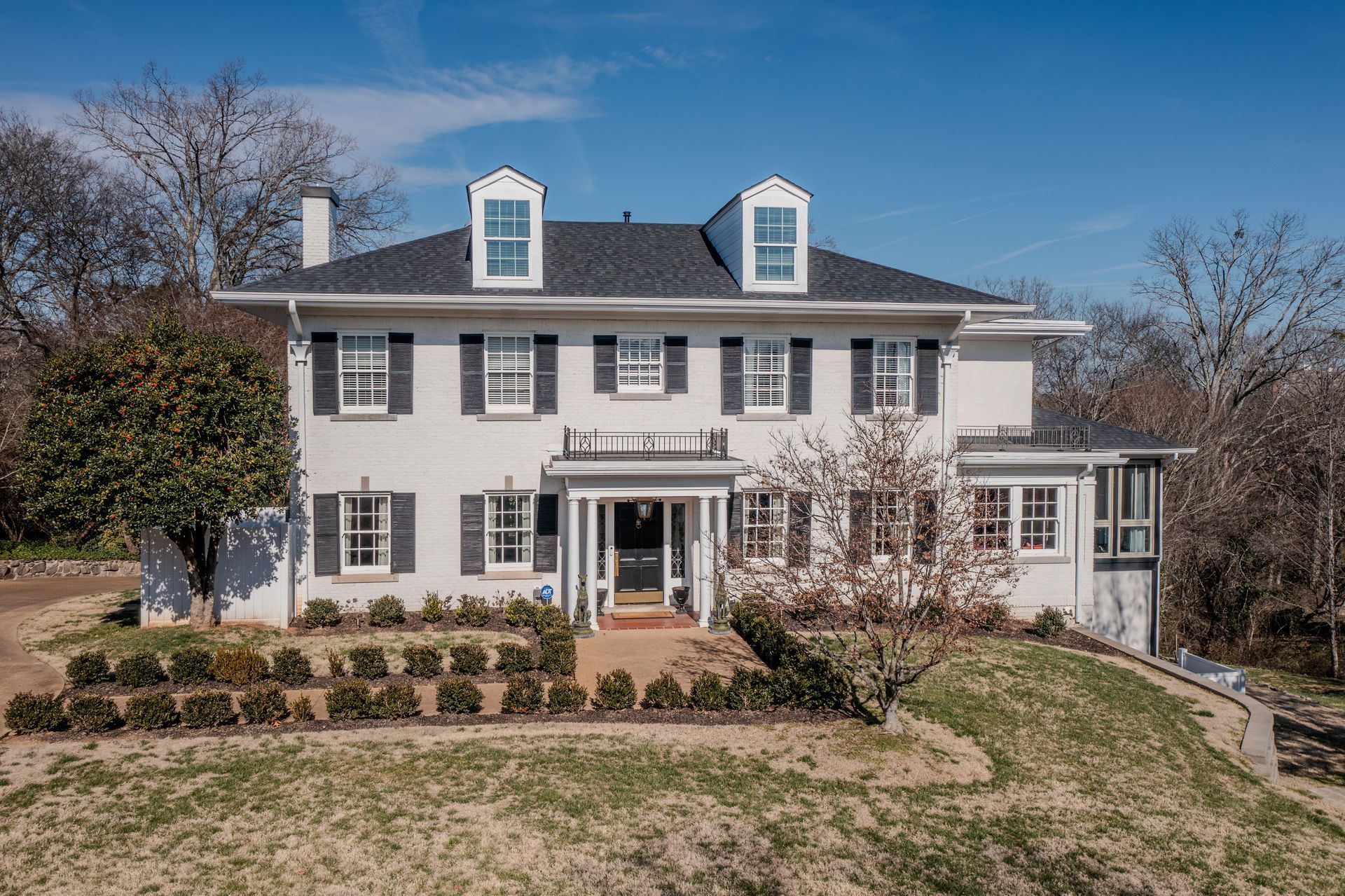 A two-story, white Colonial-style home with black shutters, dormer windows, and a front porch under a clear blue sky.
