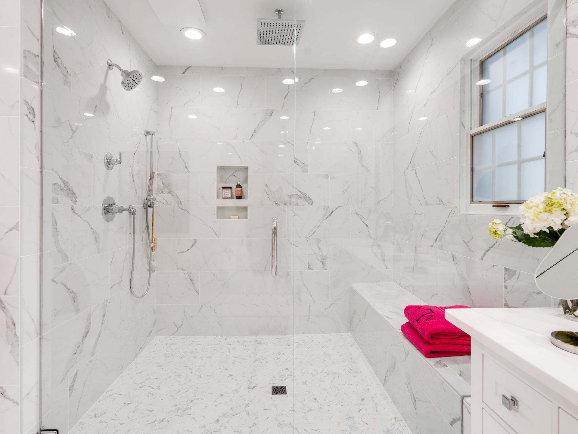 Spacious white marble shower with glass doors, built-in bench, and window; pink towels sit on bench.
