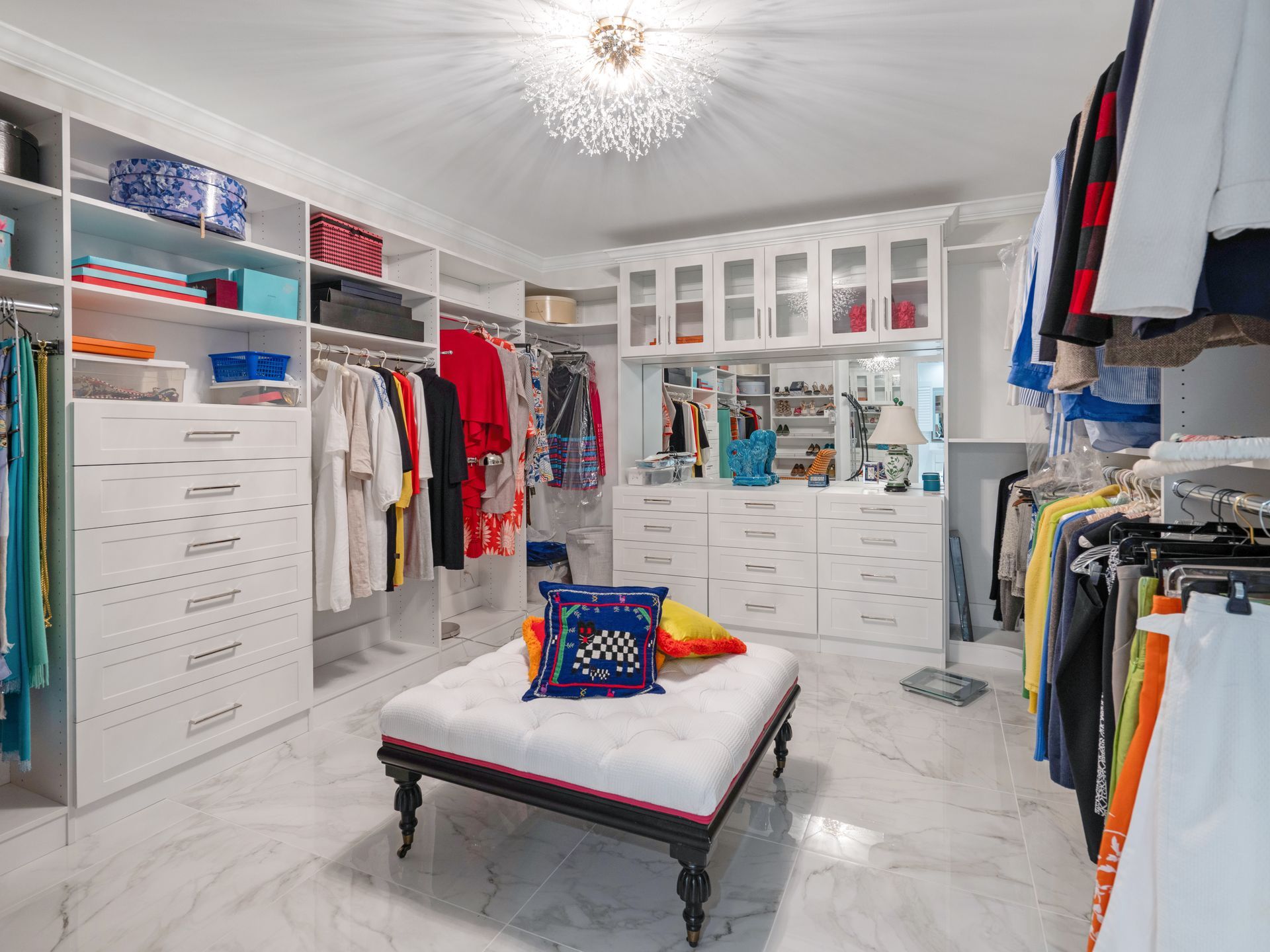 Walk-in closet with white shelves, marble floor, clothing racks, and ottoman with pillows.