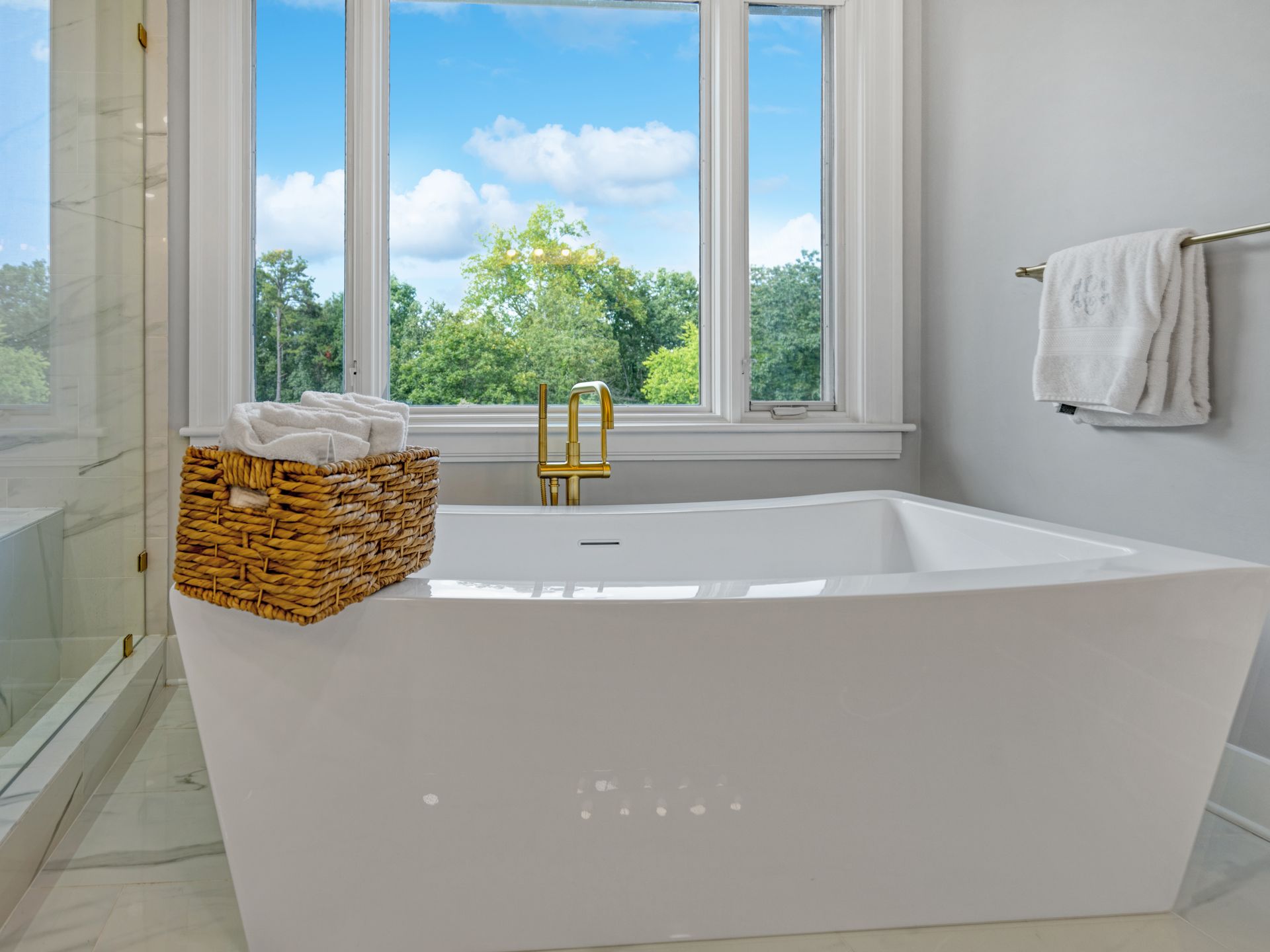 Modern bathroom with white tub, gold faucet, window with trees and blue sky.