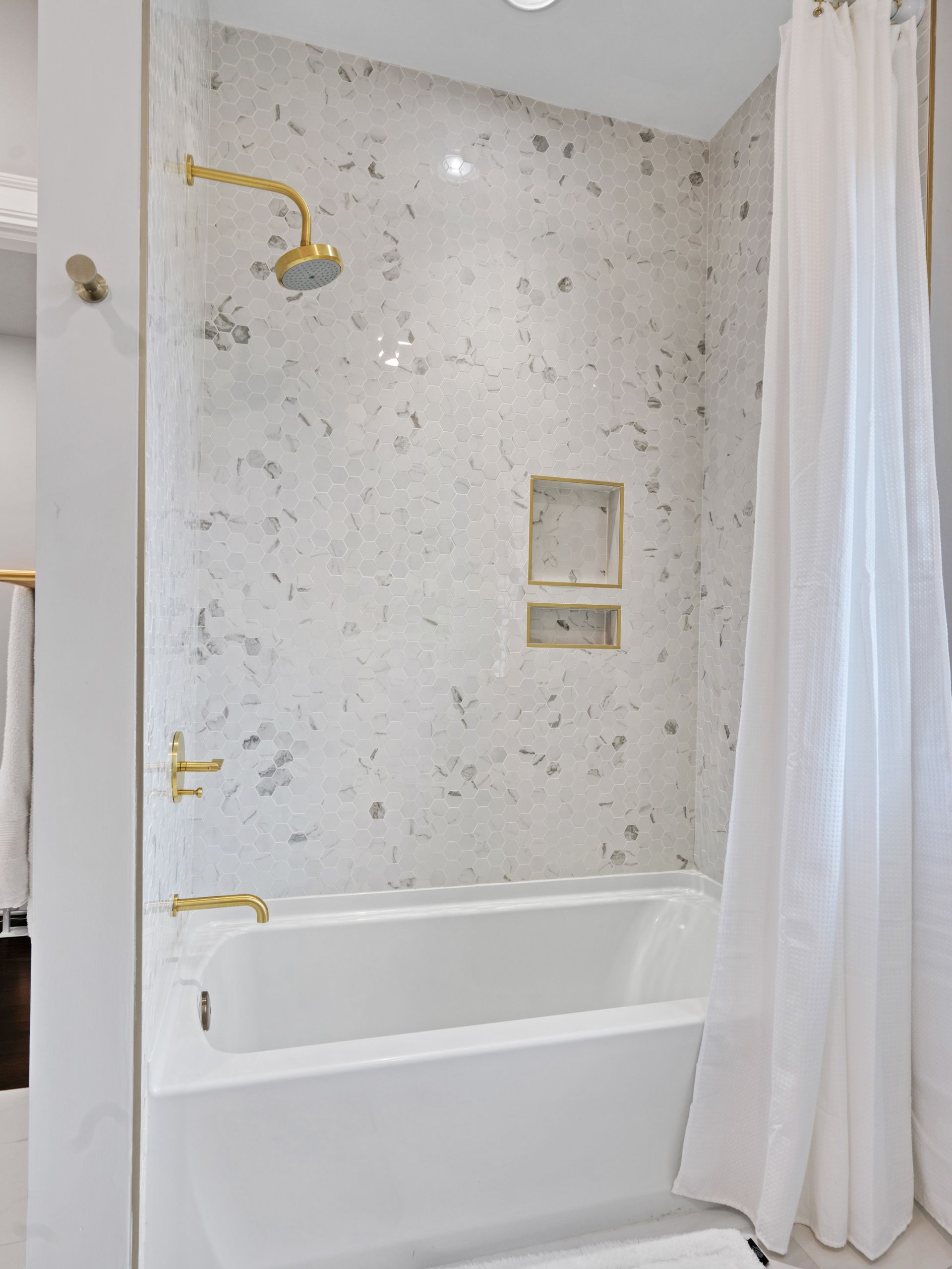 White bathroom with a bathtub, marble tile, and gold fixtures. A white shower curtain hangs to the right.