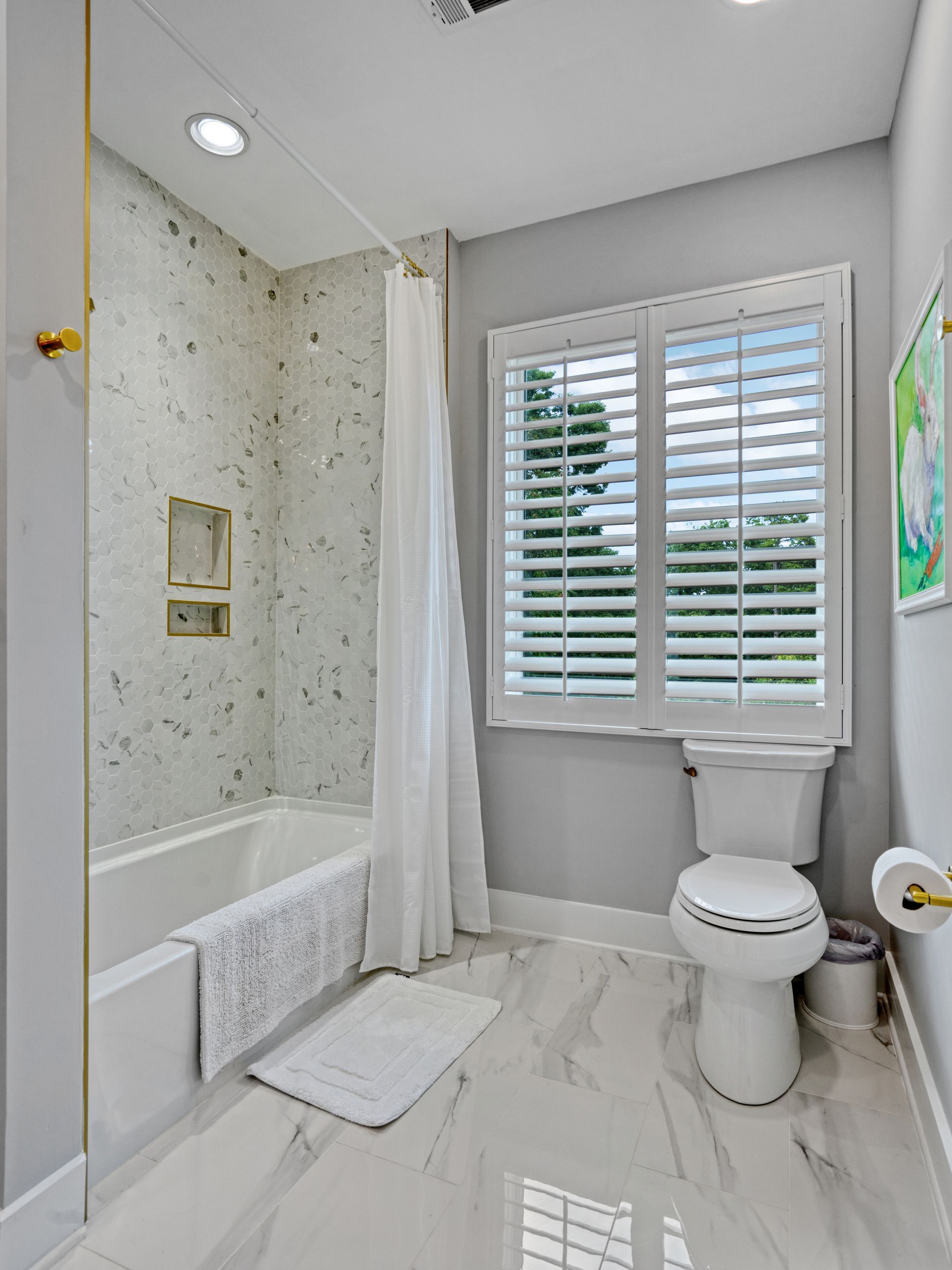Bathroom with white tile floor and walls, a bathtub, toilet, window with shutters, and a white shower curtain.