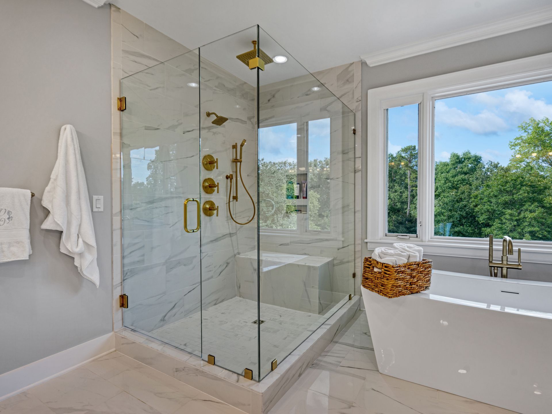 Modern bathroom with glass shower, gold fixtures, and a white bathtub next to a window overlooking trees.