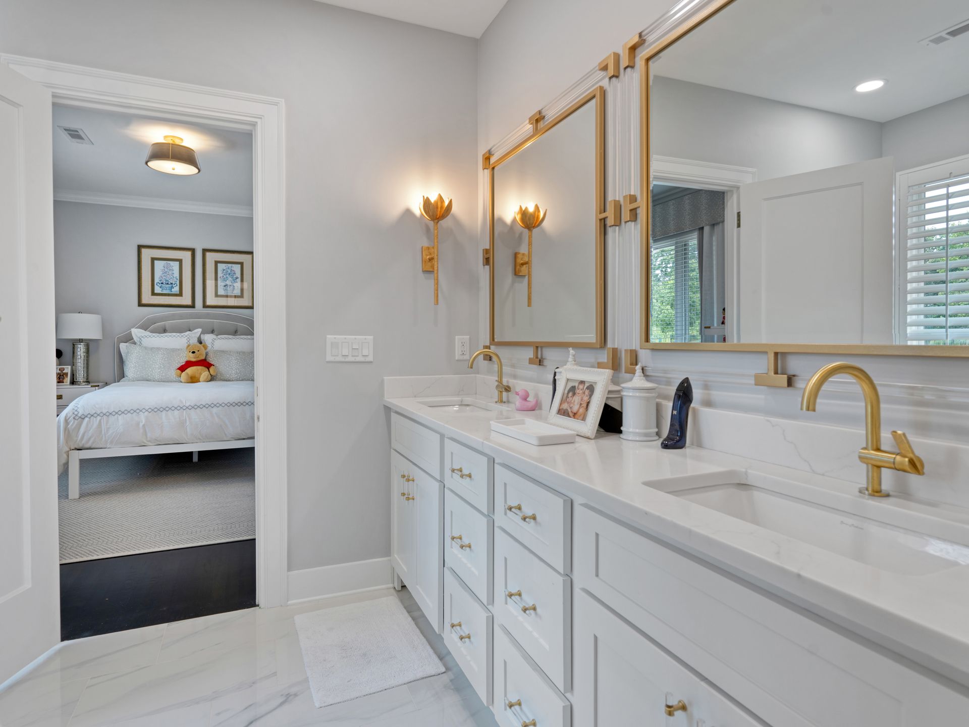 White bathroom with double vanity, gold fixtures, and a view into a bedroom.