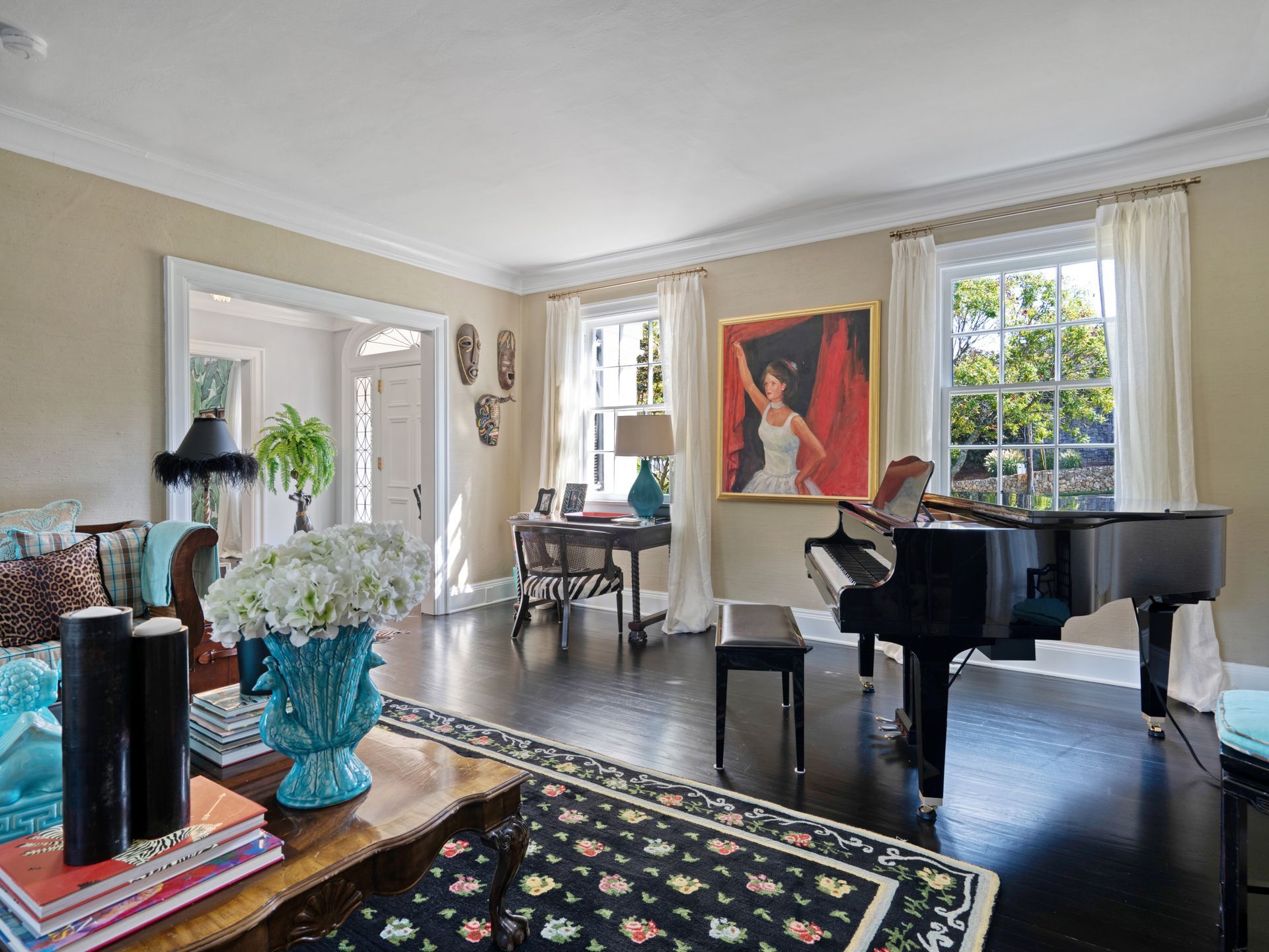 Living room with grand piano, artwork, desk, and floral arrangements.