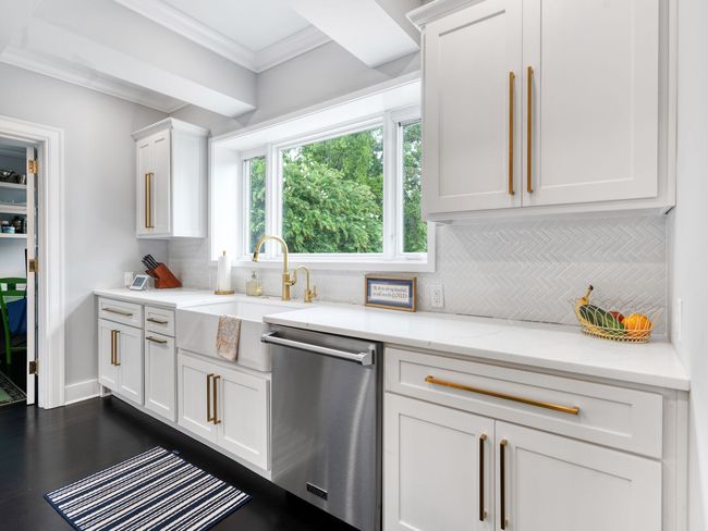 Bright white kitchen with white cabinets, gold hardware, stainless steel appliances, and a window.