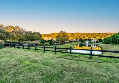 Green grassy field with black fence, a pond with a fountain, and houses in the distance under a blue sky.
