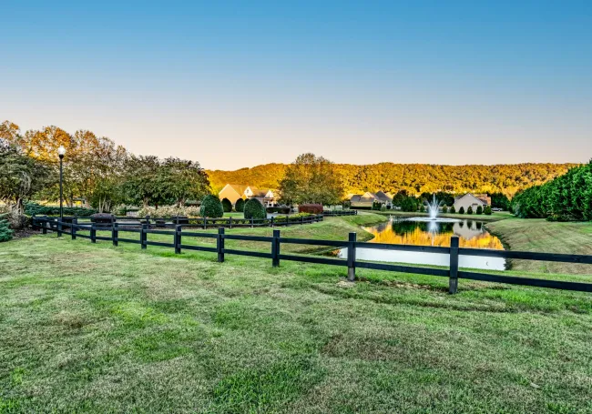 Green grassy field with black fence, a pond with a fountain, and houses in the distance under a blue sky.