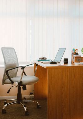 An office setup featuring a light-colored ergonomic mesh chair positioned at a wooden desk with a laptop and supplies.