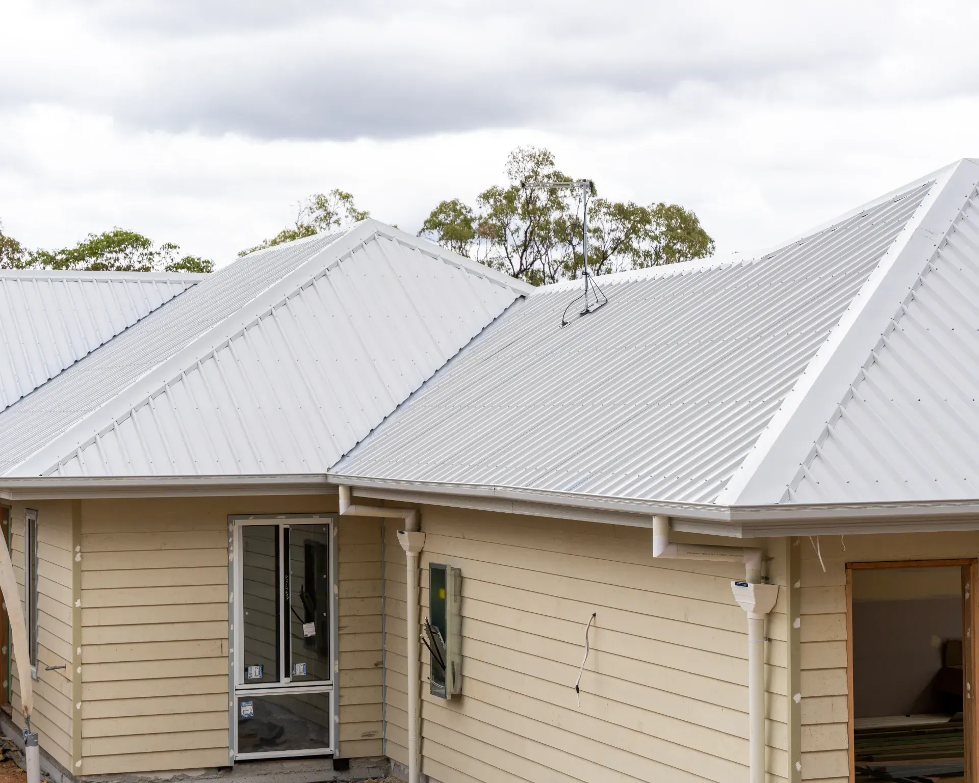 Exterior view of a light-colored, unfinished house with light grey corrugated metal roofing and cream-colored siding.