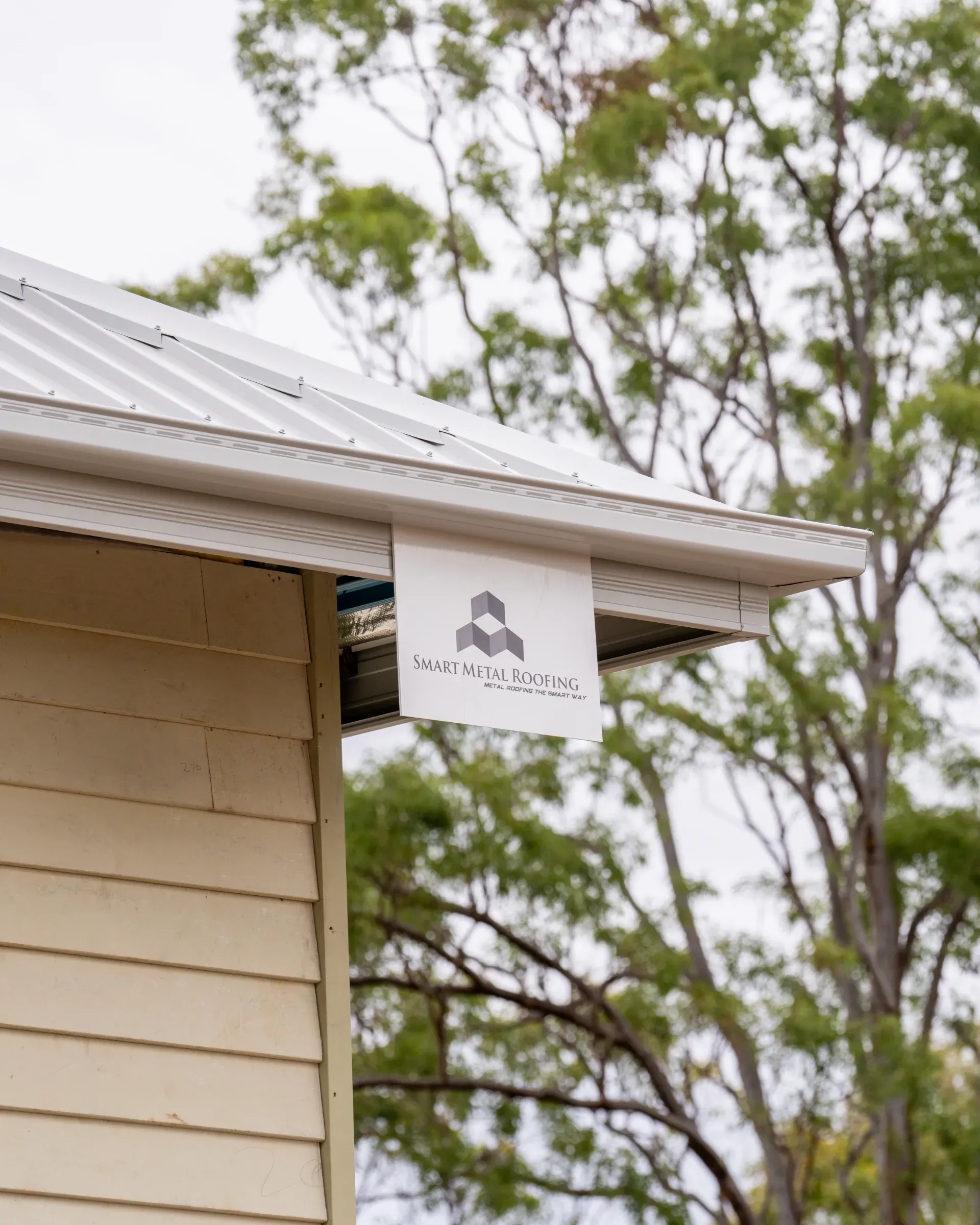 A white sign with a gray abstract logo hangs from the corrugated metal gutter of a light-yellow weatherboard house.