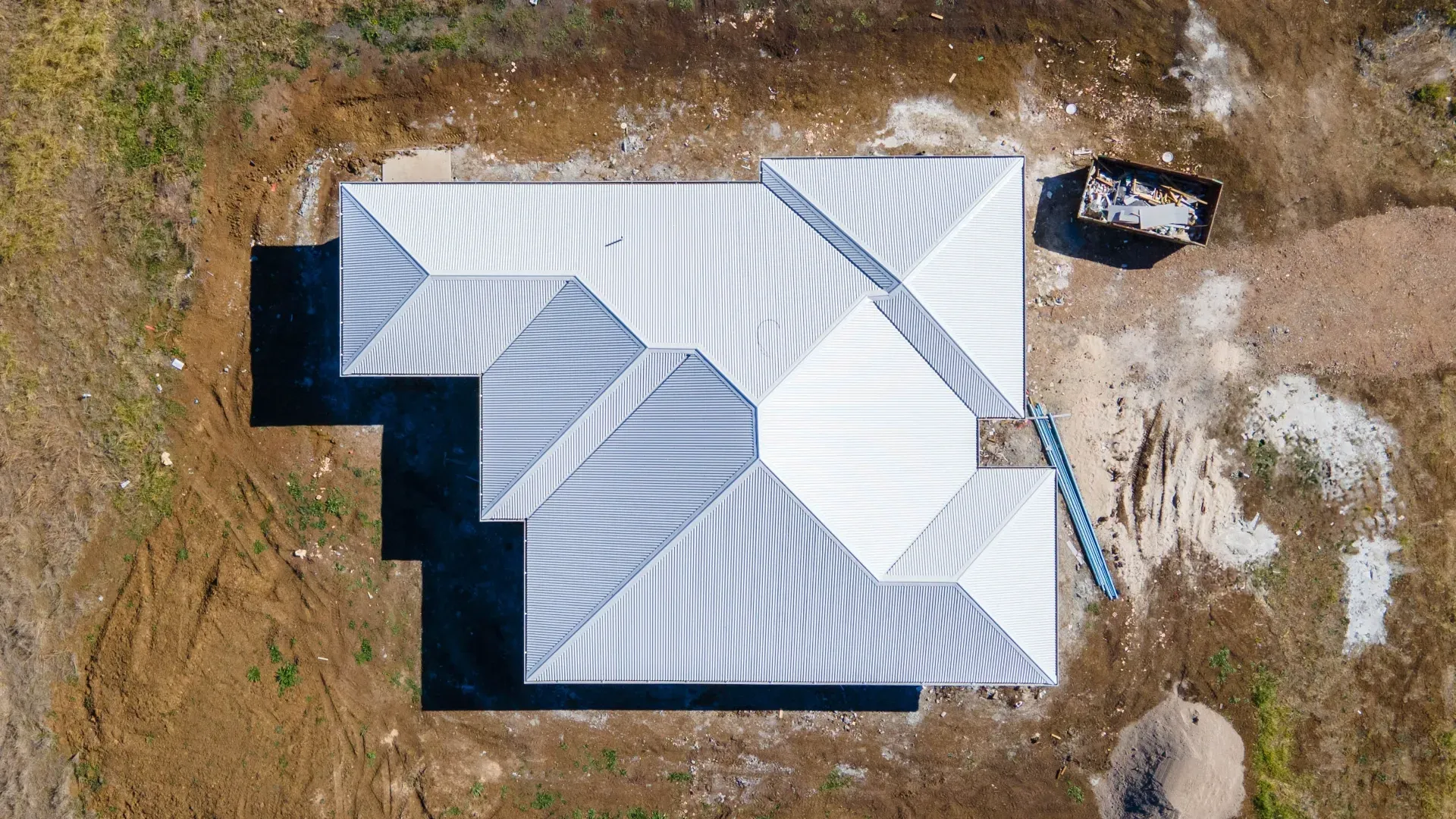 An aerial view of a light-colored, multi-pitched metal roof on a house under construction amidst bare dirt and grass.