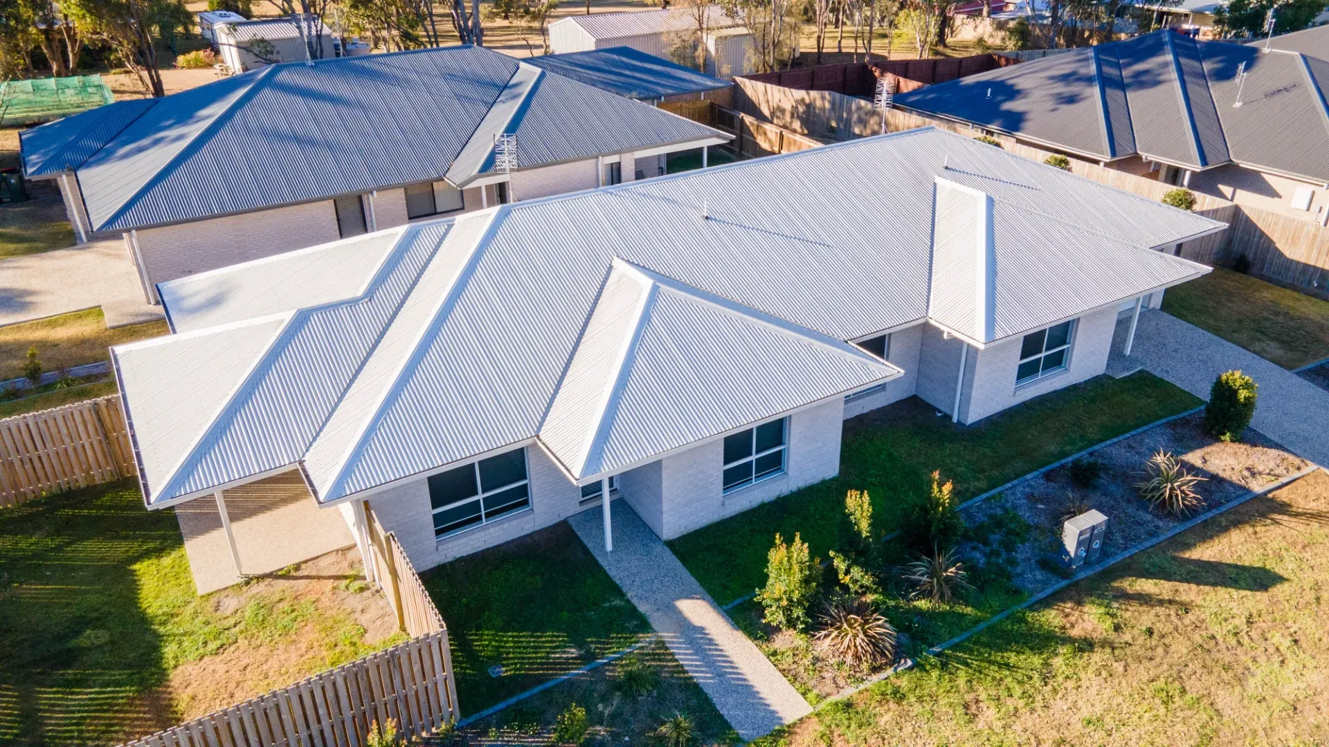 An elevated, high-angle view of a modern suburban house with a gray corrugated metal roof, white walls, and a small yard.