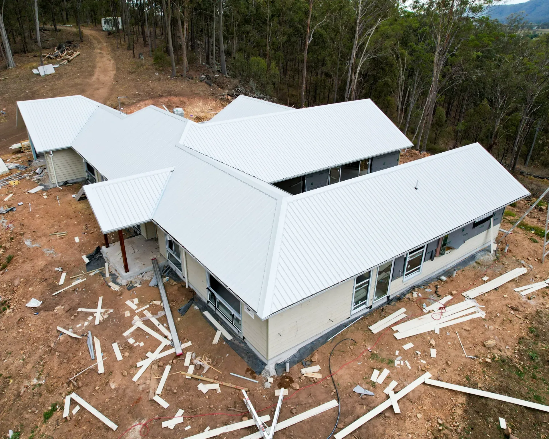 An aerial view of a new, single-story house with a light-colored metal roof under construction in a wooded area.