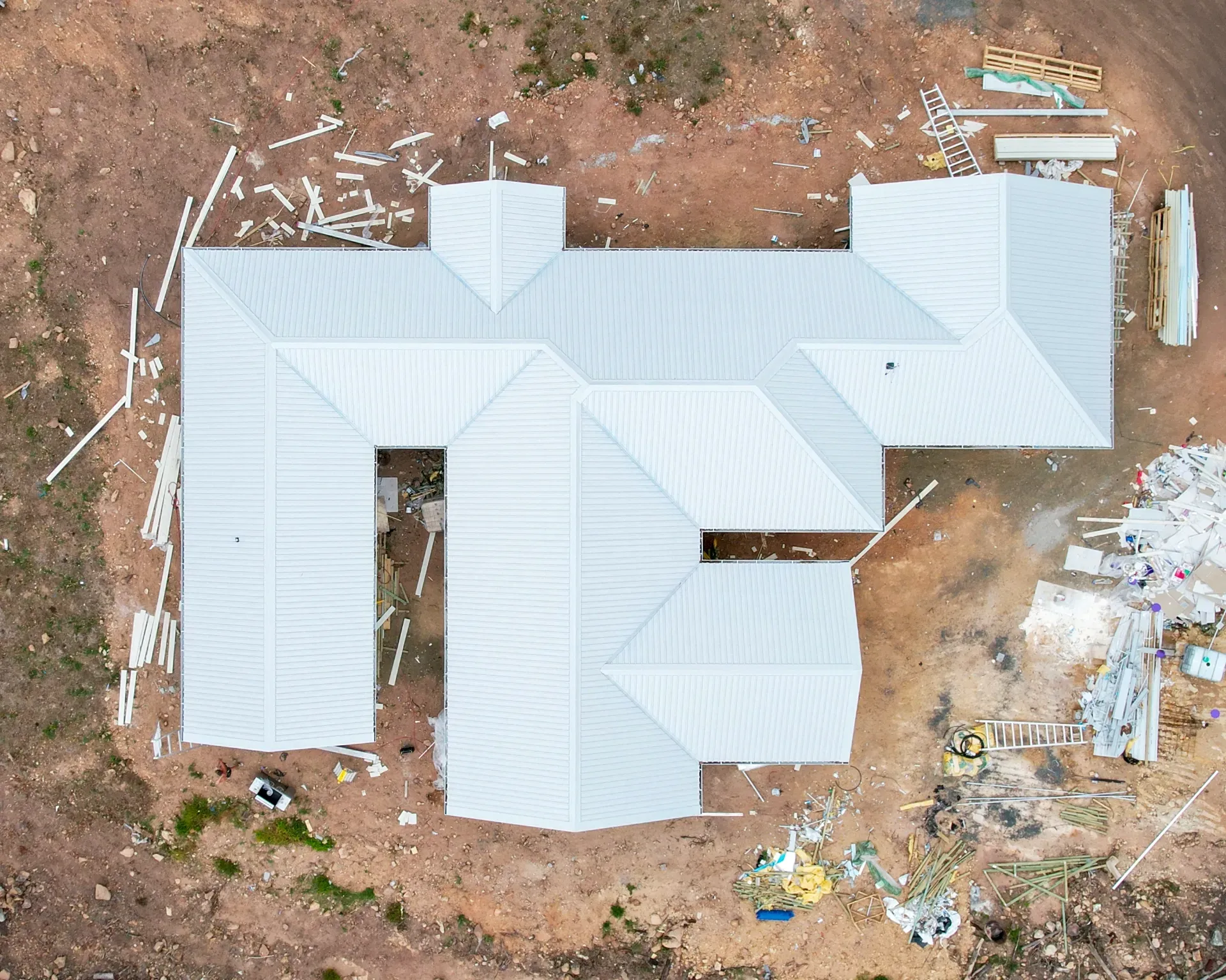 An aerial view of a light-colored, multi-gabled house under construction on a dirt lot with scattered building debris.