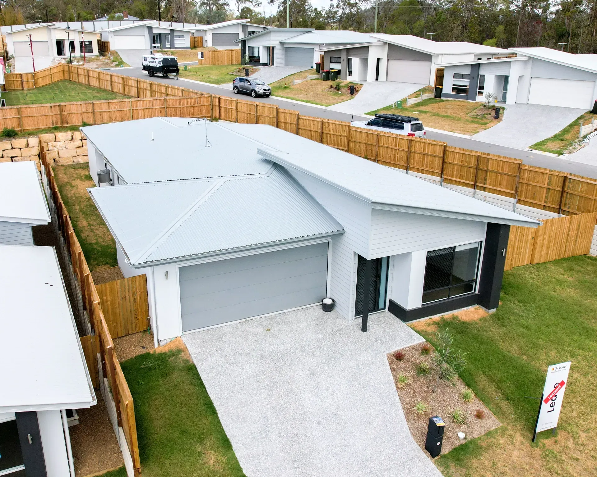 An aerial view of a modern light-gray house with a flat roof, gravel driveway, and wood fence in a suburban neighborhood.