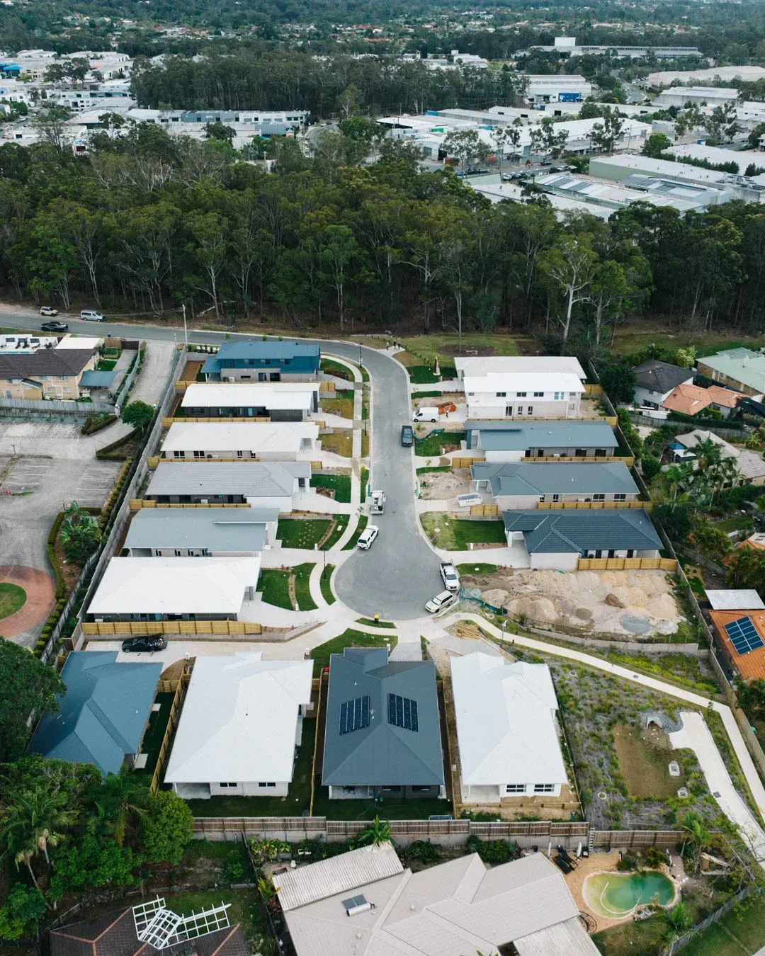 An aerial view of a new suburban residential street with rows of modern houses and driveways surrounded by green trees.