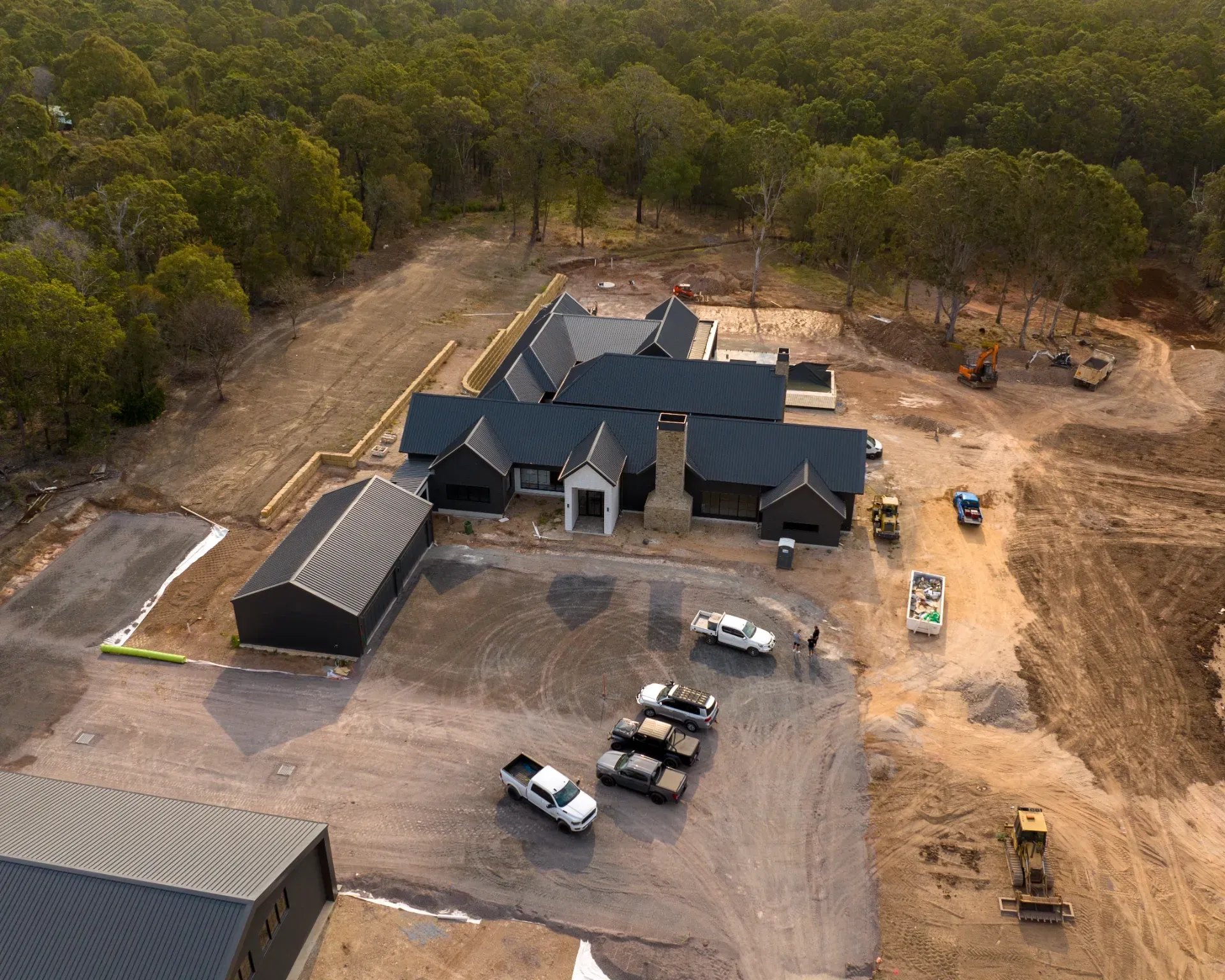 An aerial view of a modern black farmhouse-style home under construction, surrounded by woods with trucks in the driveway.