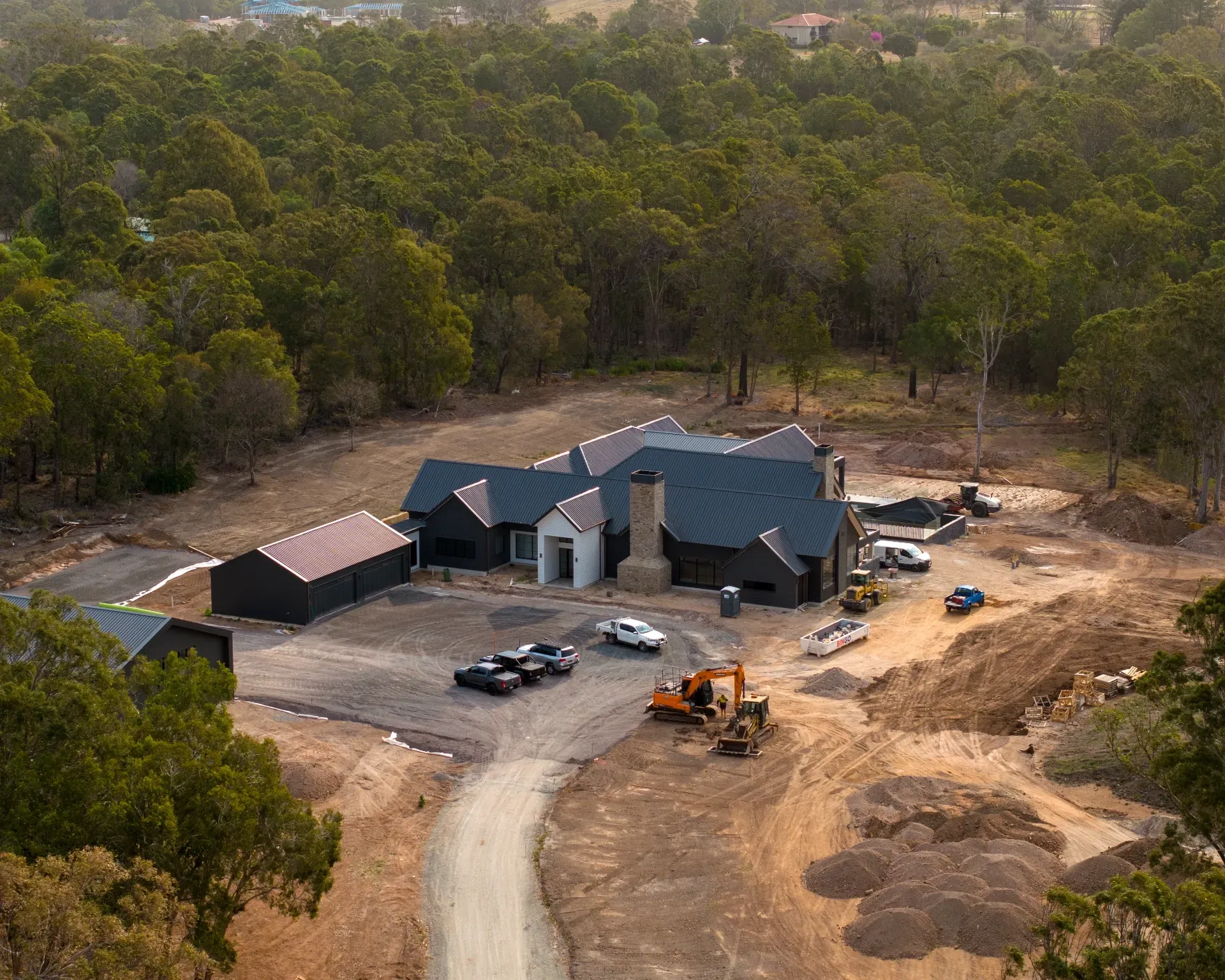 An aerial view of a dark-colored, modern house under construction, surrounded by a dense forest and a dirt driveway.