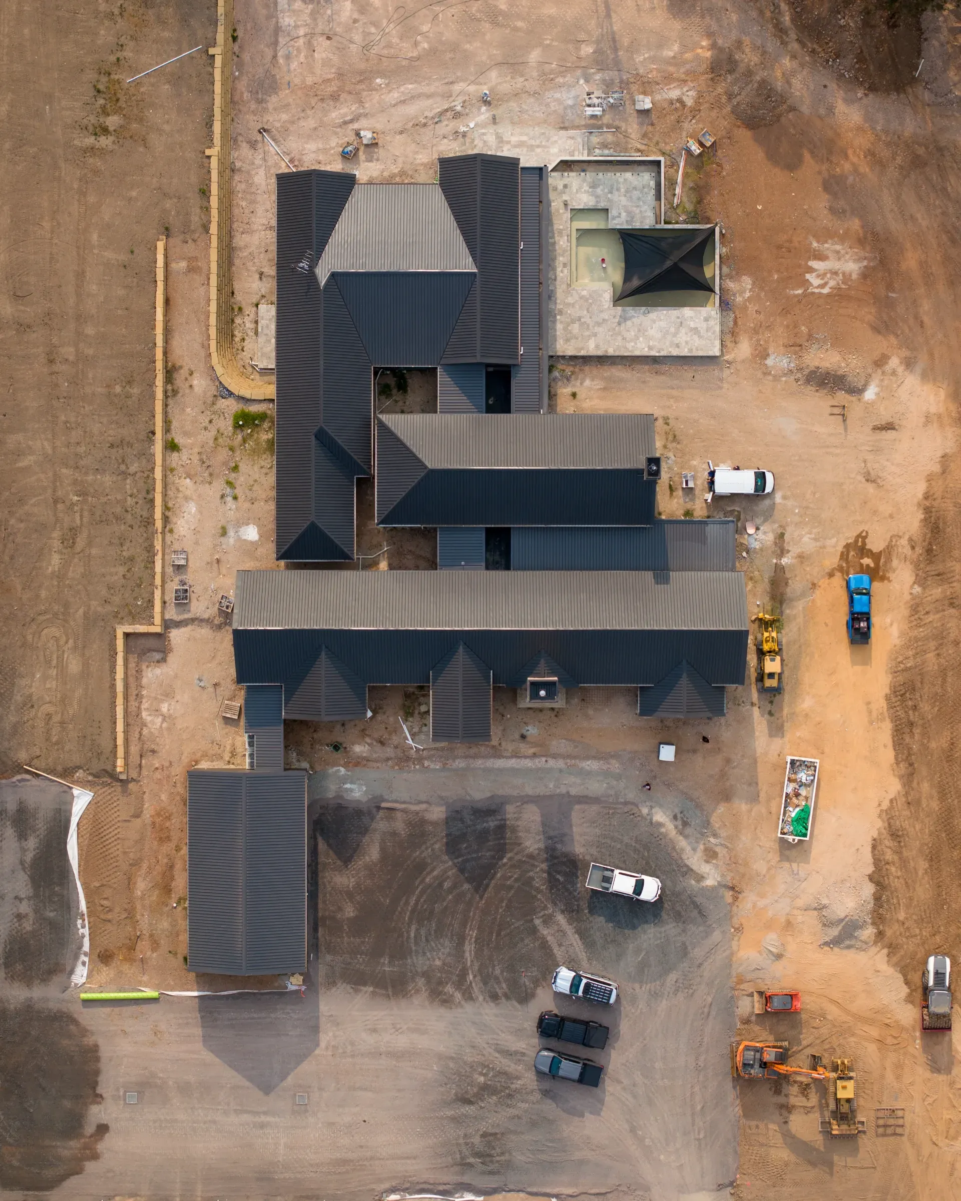 An aerial view of a construction site featuring a newly framed house with a dark metal roof, vehicles, and dirt grounds.