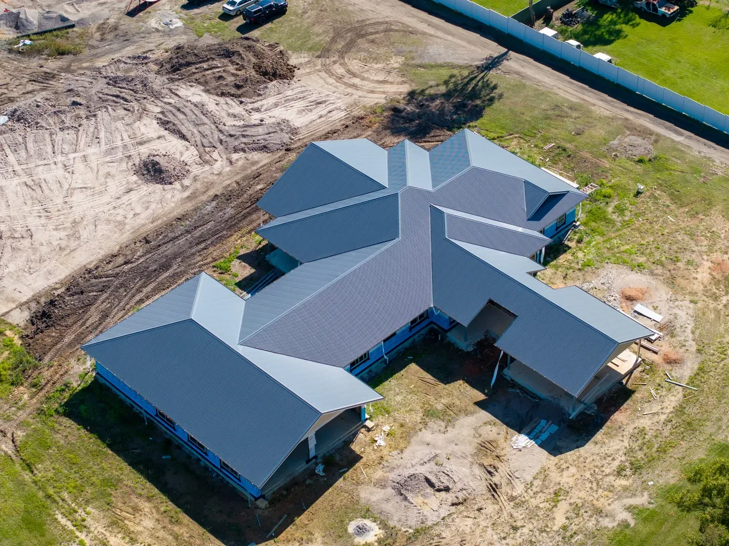An aerial view of a new residential home under construction, featuring a complex grey roof and surrounding dirt site.