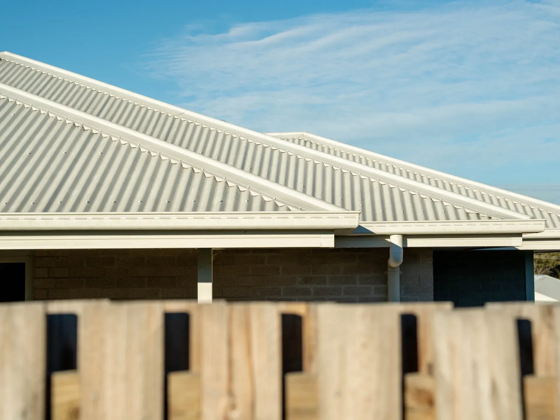 A light-colored corrugated metal roof with white gutters, seen behind a blurred wooden fence against a clear blue sky.