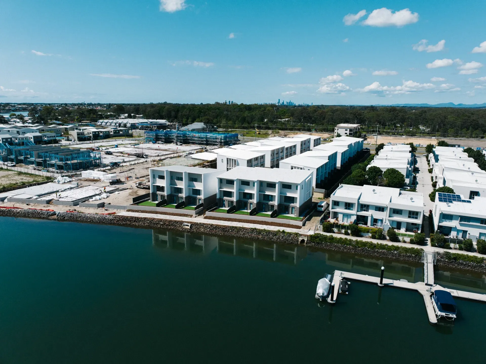 Modern townhouses by water with boat docks, construction in background, sunny day. — Smart Metal Roofing in Coomera, QLD