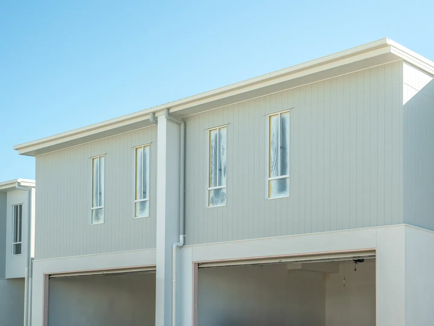 Two-story modern townhouses with light gray exterior walls, garage doors, and tall, narrow windows against a clear blue sky. — Smart Metal Roofing in Coomera, QLD