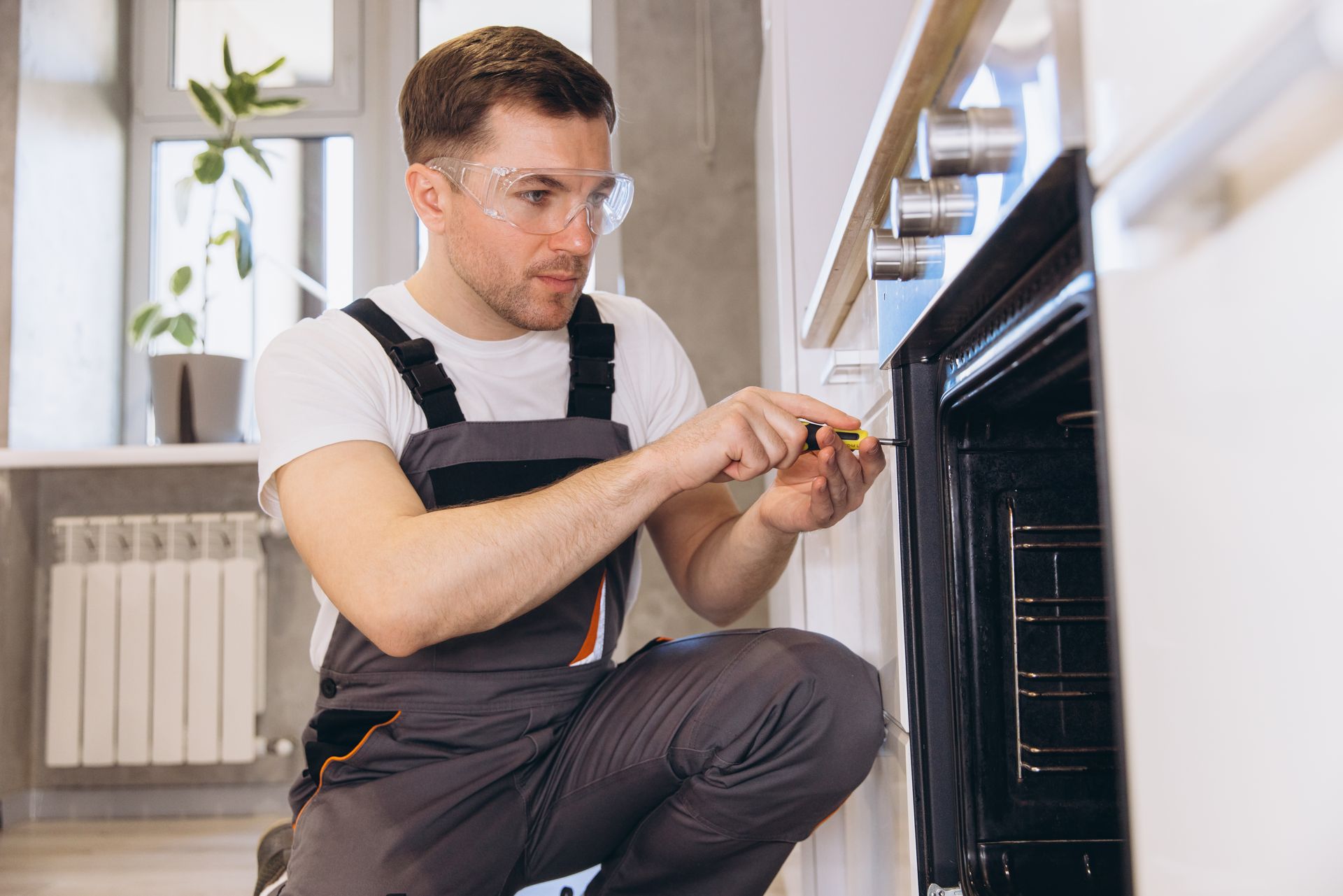 Technician kneeling by an oven, using a screwdriver to make repairs in a kitchen.
