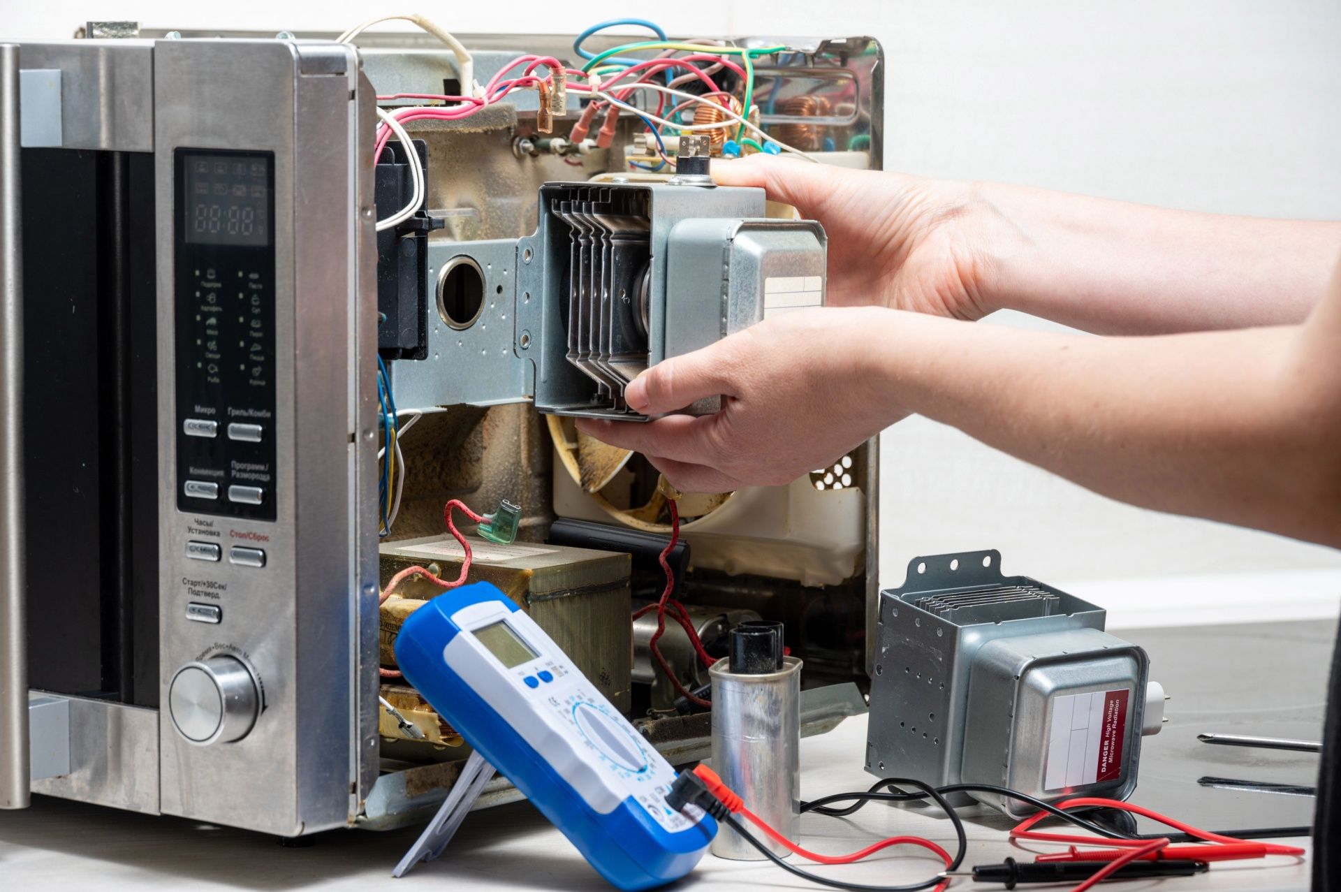 Hands removing a microwave’s internal component, with wires and tools exposed on a workbench.